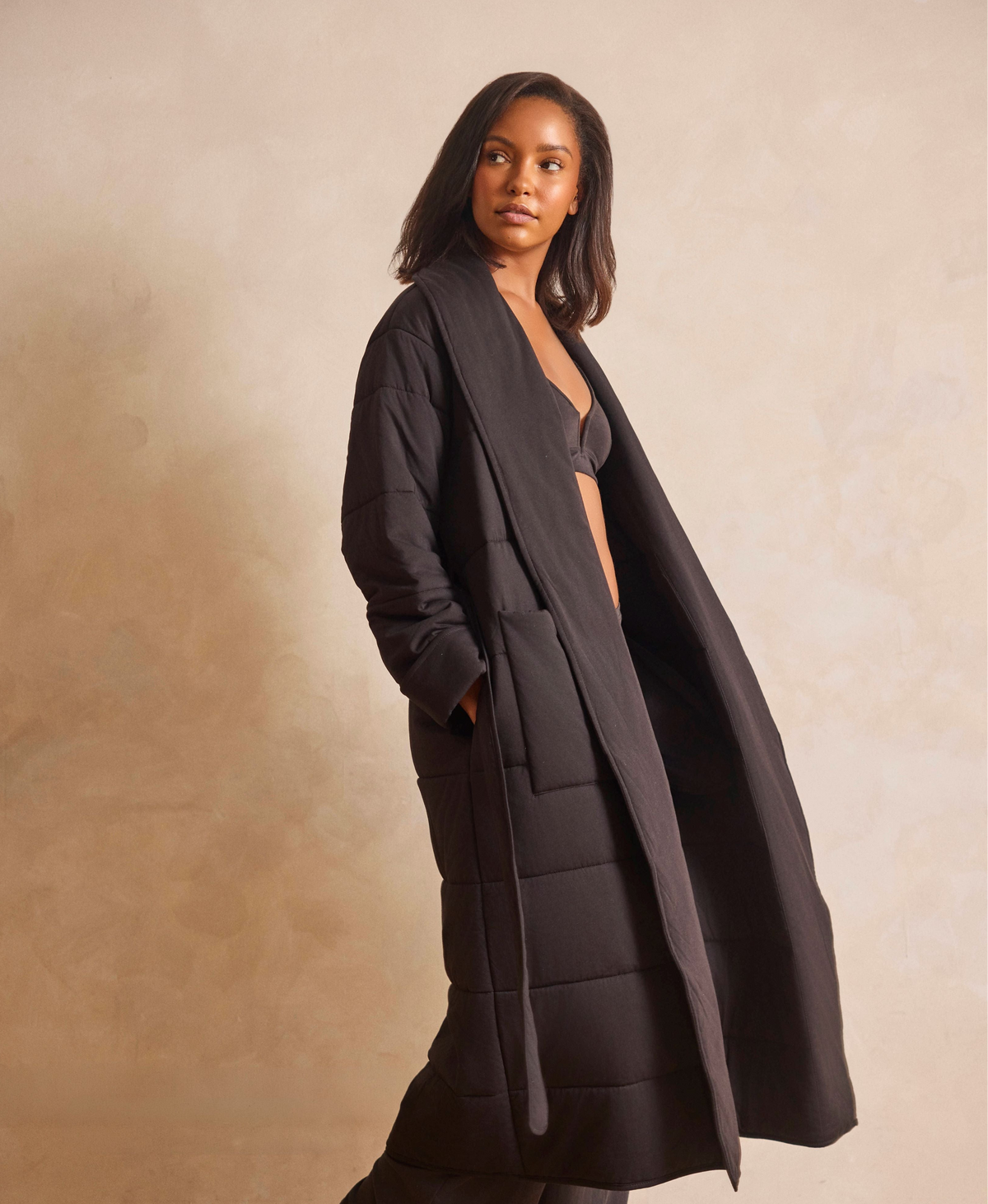 A woman modeling a long black quilted puffer coat with a shawl collar and front buttons, posed against a warm beige textured studio backdrop.
