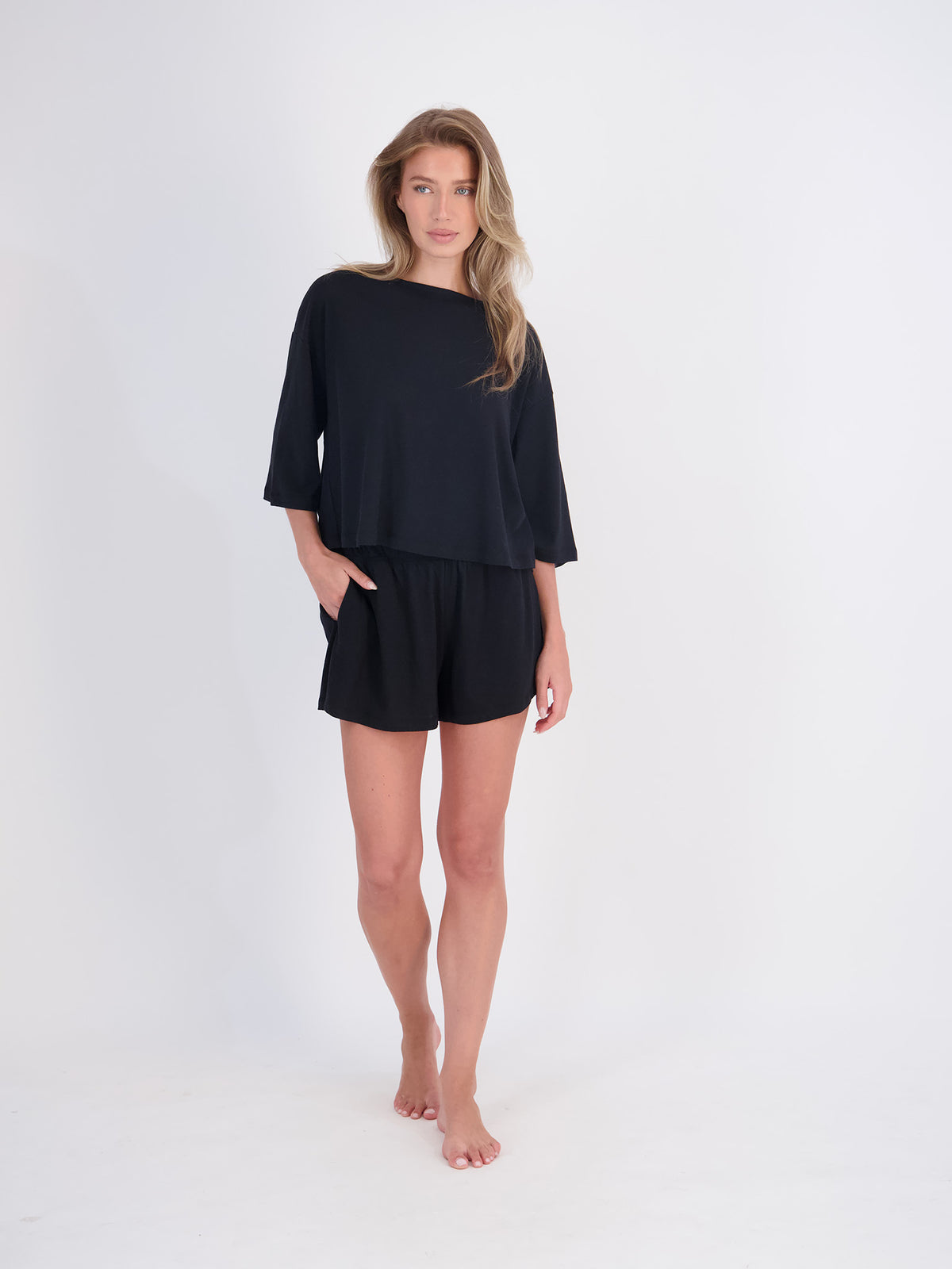A woman modeling a black two-piece lounge set consisting of a short-sleeve top and matching shorts, standing against a plain white studio background.