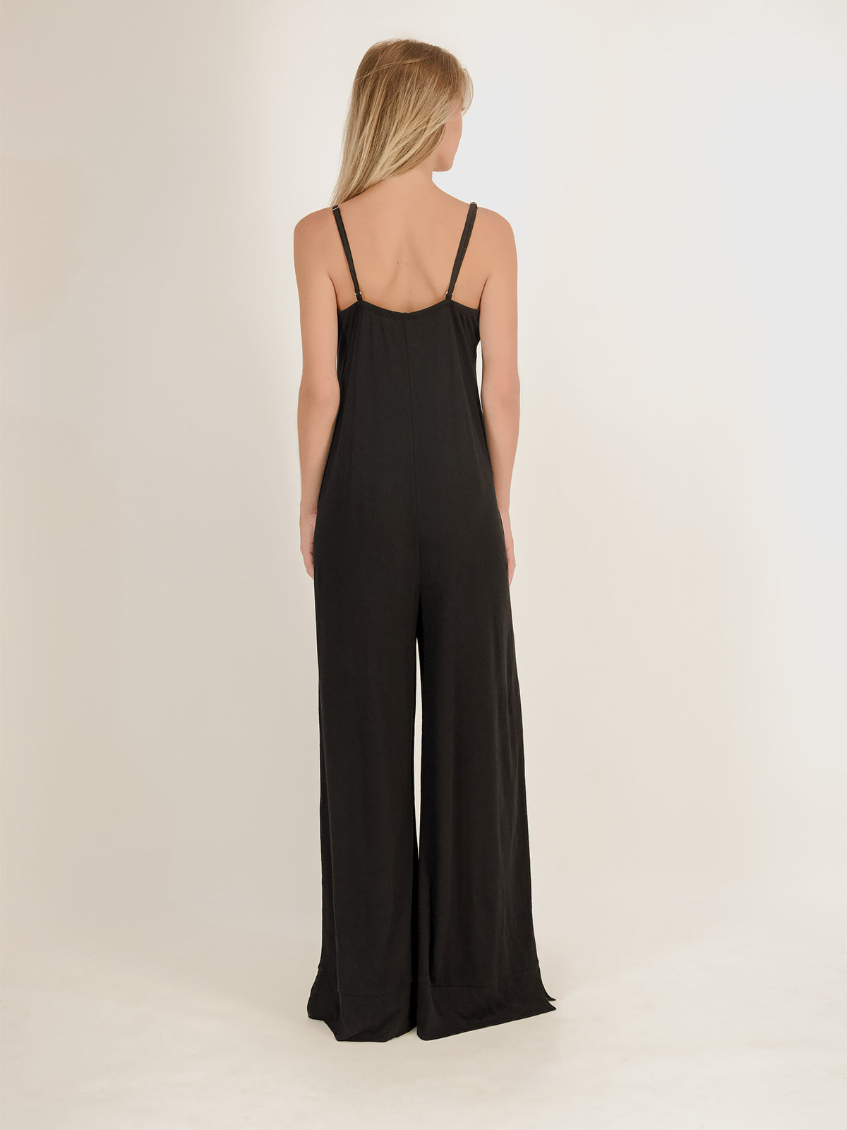 A woman viewed from the back wearing a black sleeveless wide-leg jumpsuit with thin shoulder straps, standing against a plain light studio background.