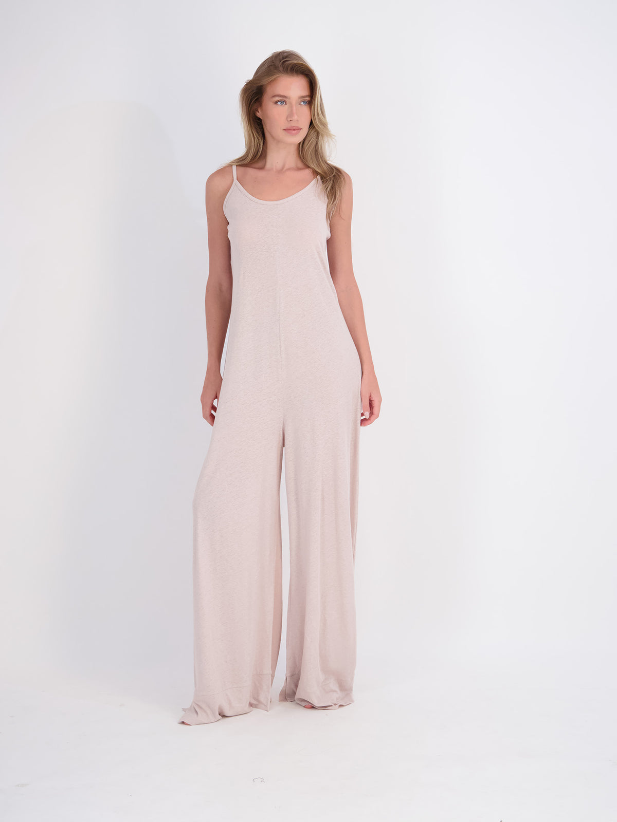 A woman modeling a light mushroom beige sleeveless jumpsuit with wide legs, standing against a plain white studio background.