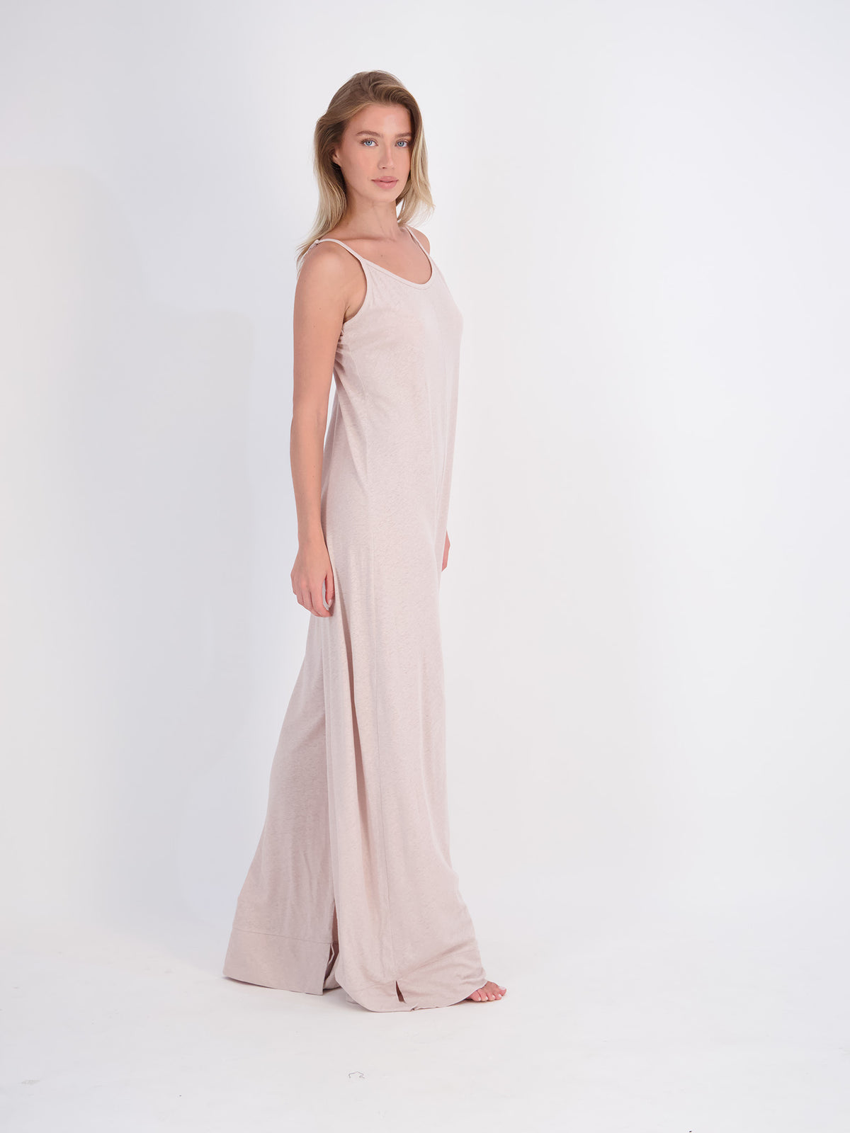 A woman modeling a sleeveless, floor-length jumpsuit in a soft mushroom beige color, standing against a seamless white studio background.