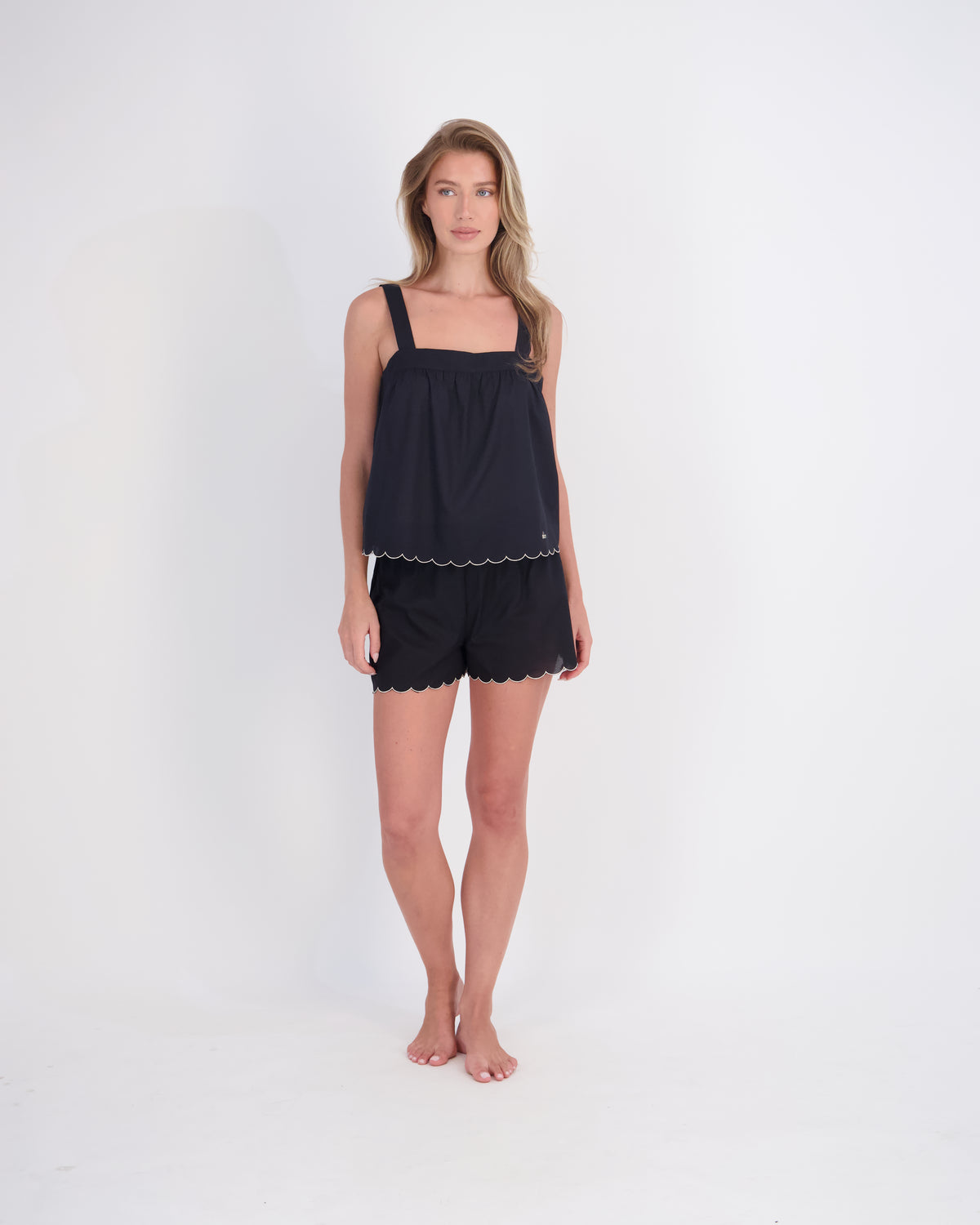 A woman modeling a black two-piece lounge set consisting of a sleeveless top with thin straps and scalloped edges, paired with matching black shorts, against a plain white studio background.